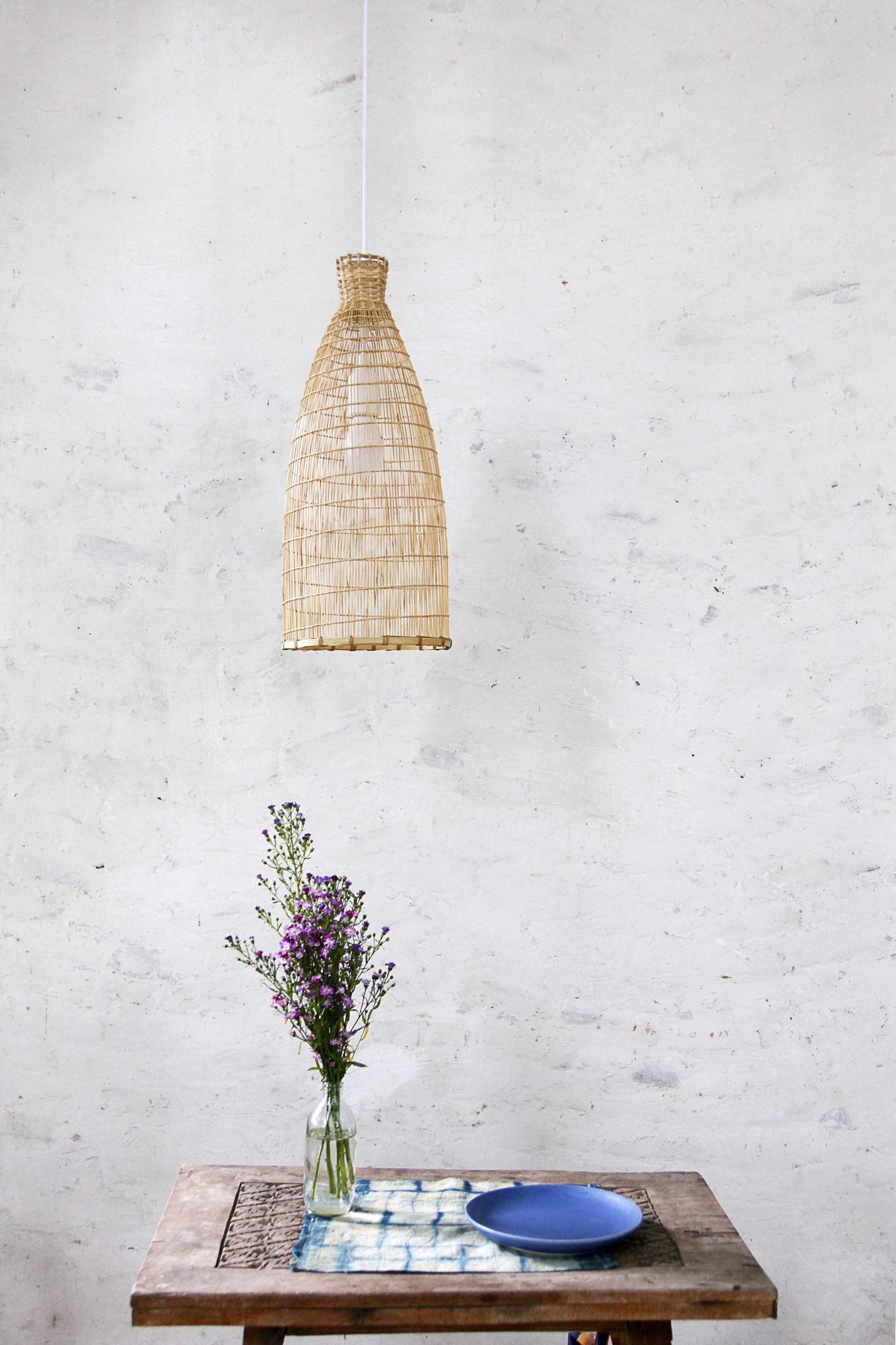 Boho Bamboo pendant light hanging above a wooden table with a vase of flowers and a plate on a white brick wall background.