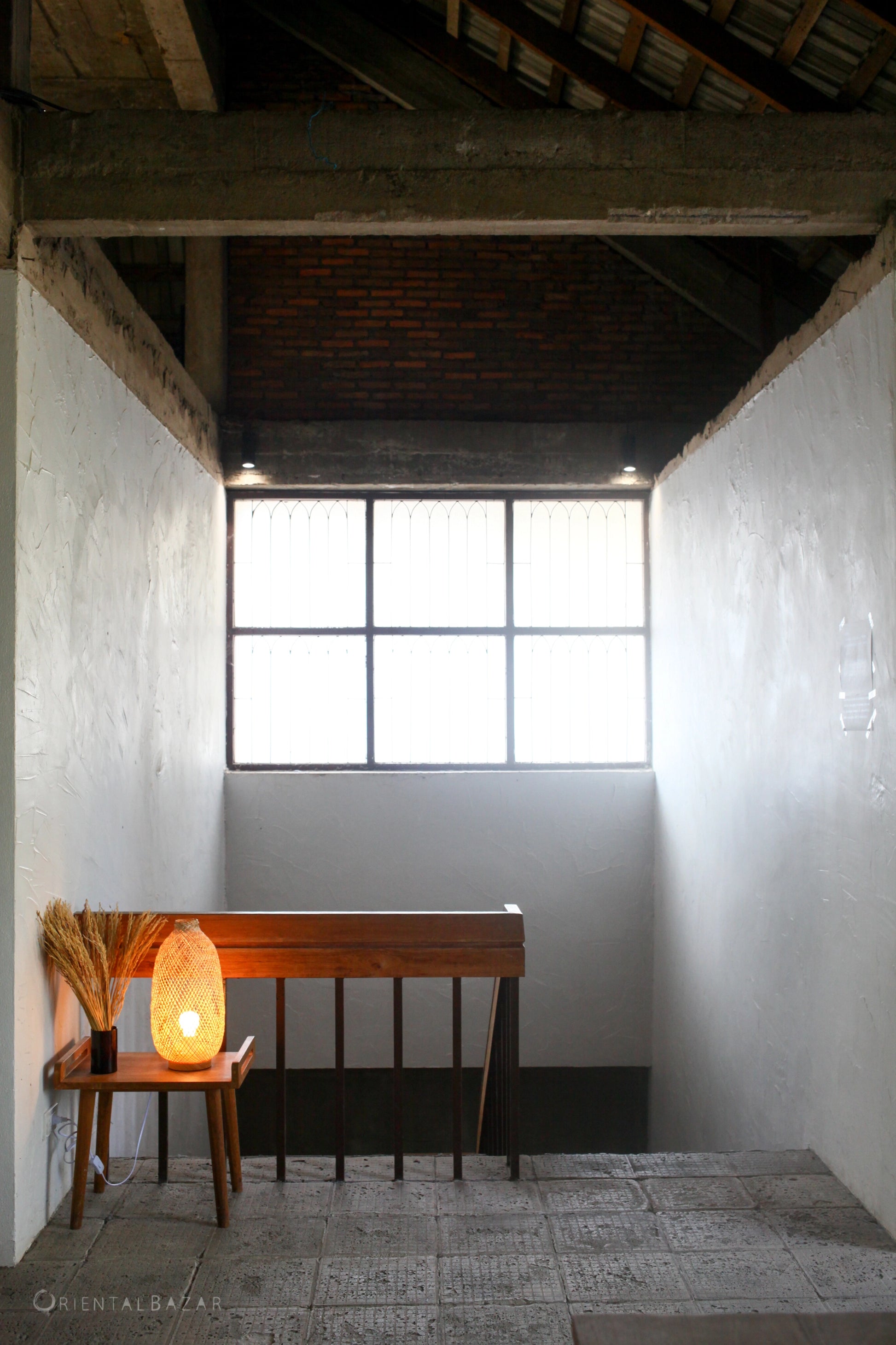 Bamboo standing lamp on a small wooden bench against a white wall with a window above, in a room with exposed wooden beams.