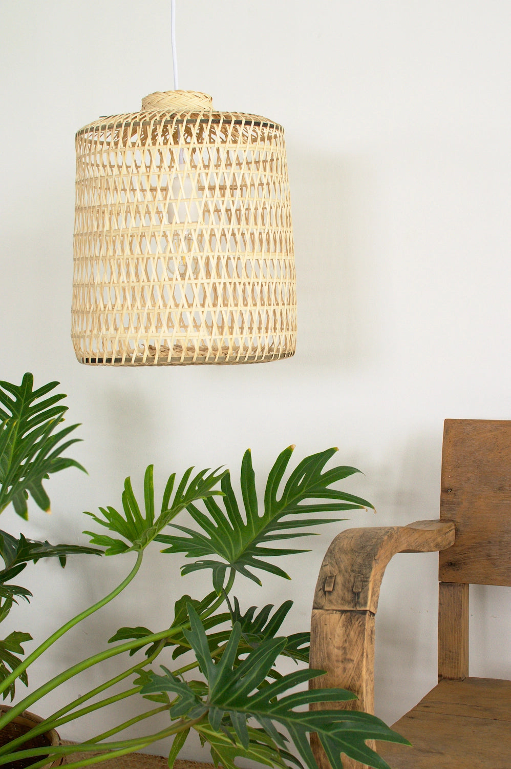 Woven bamboo pendant light hanging above a plant and wooden chair against a white wall.