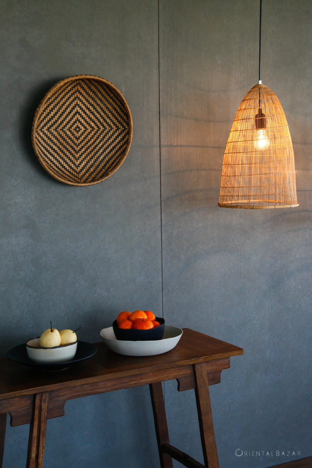 Bamboo pendant lamp hanging from ceiling in a scenario with a decorative bamboo basketry hanging on the wall and a wooden table with ceramic plates with fruits on it
