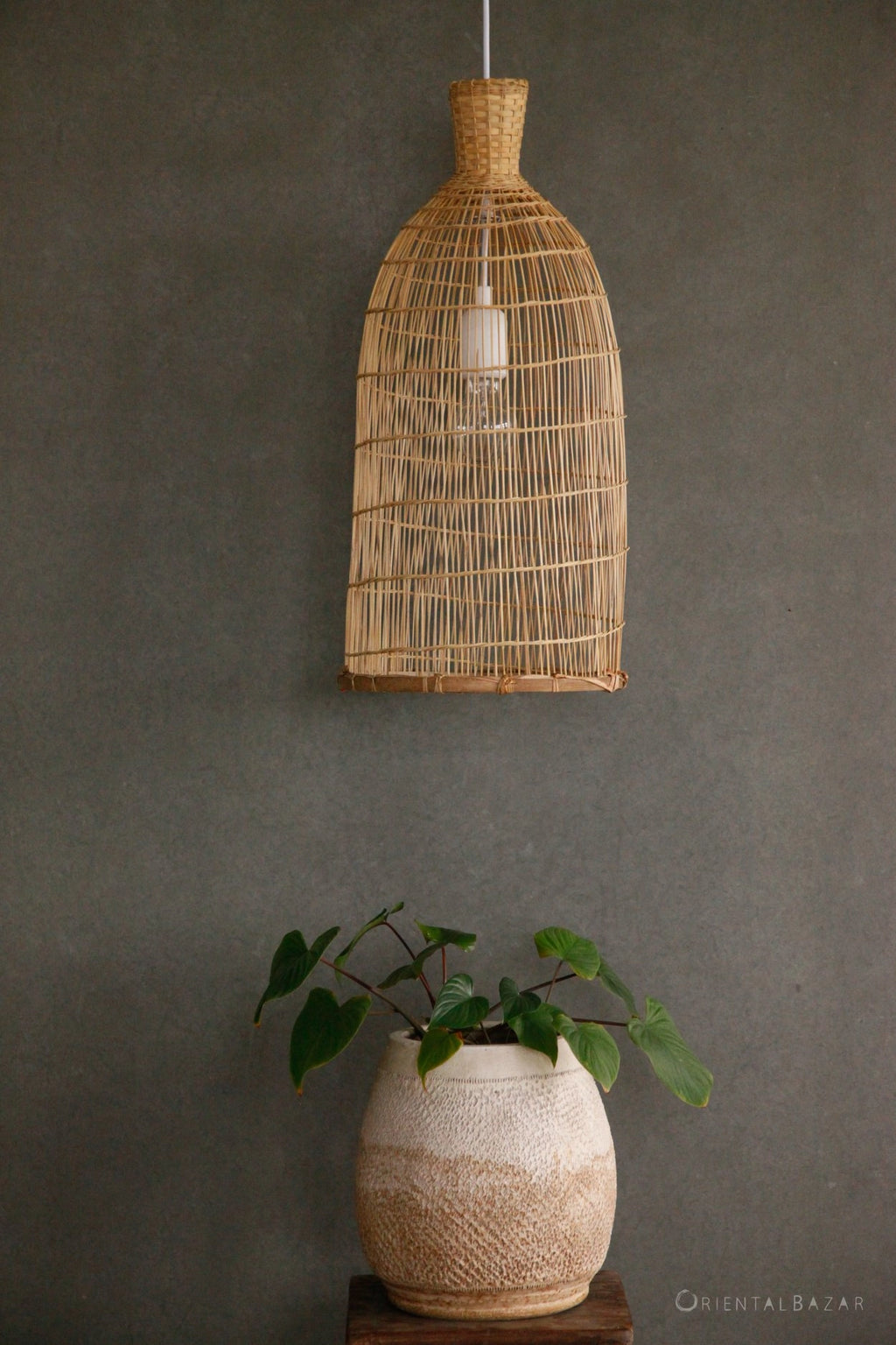 Woven bamboo pendant light fixture hanging above a plant in a textured vase against a dark gray wall.