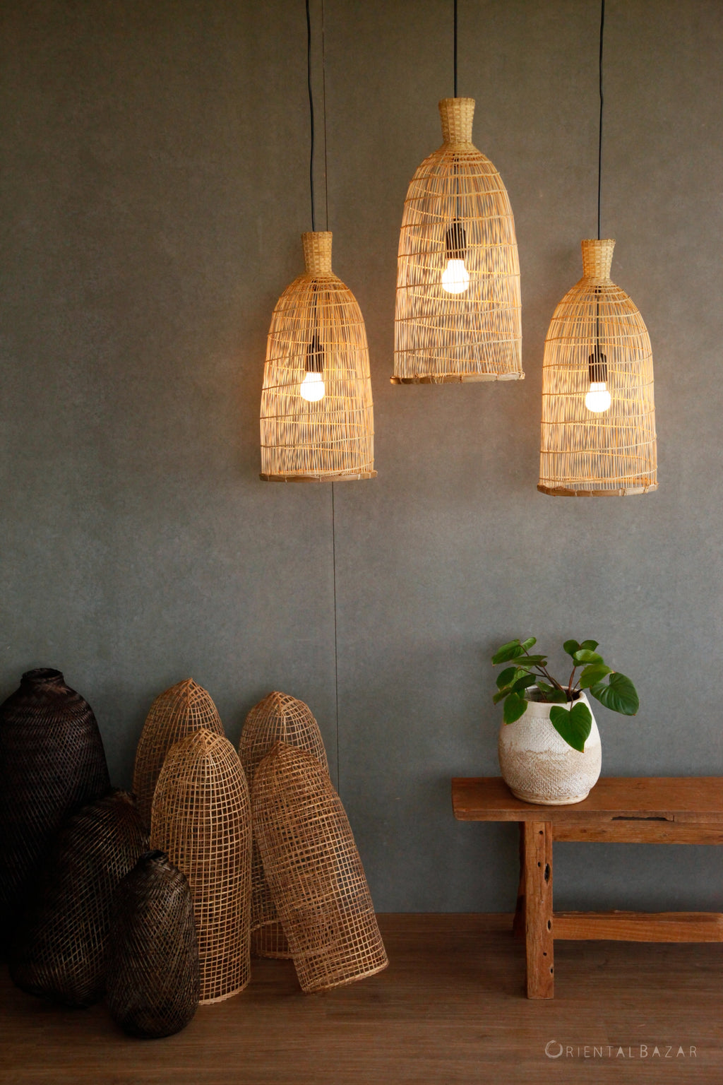 Woven pendant lights hanging against a gray wall with a wooden bench and plant and decorative bamboo baskets on the ground