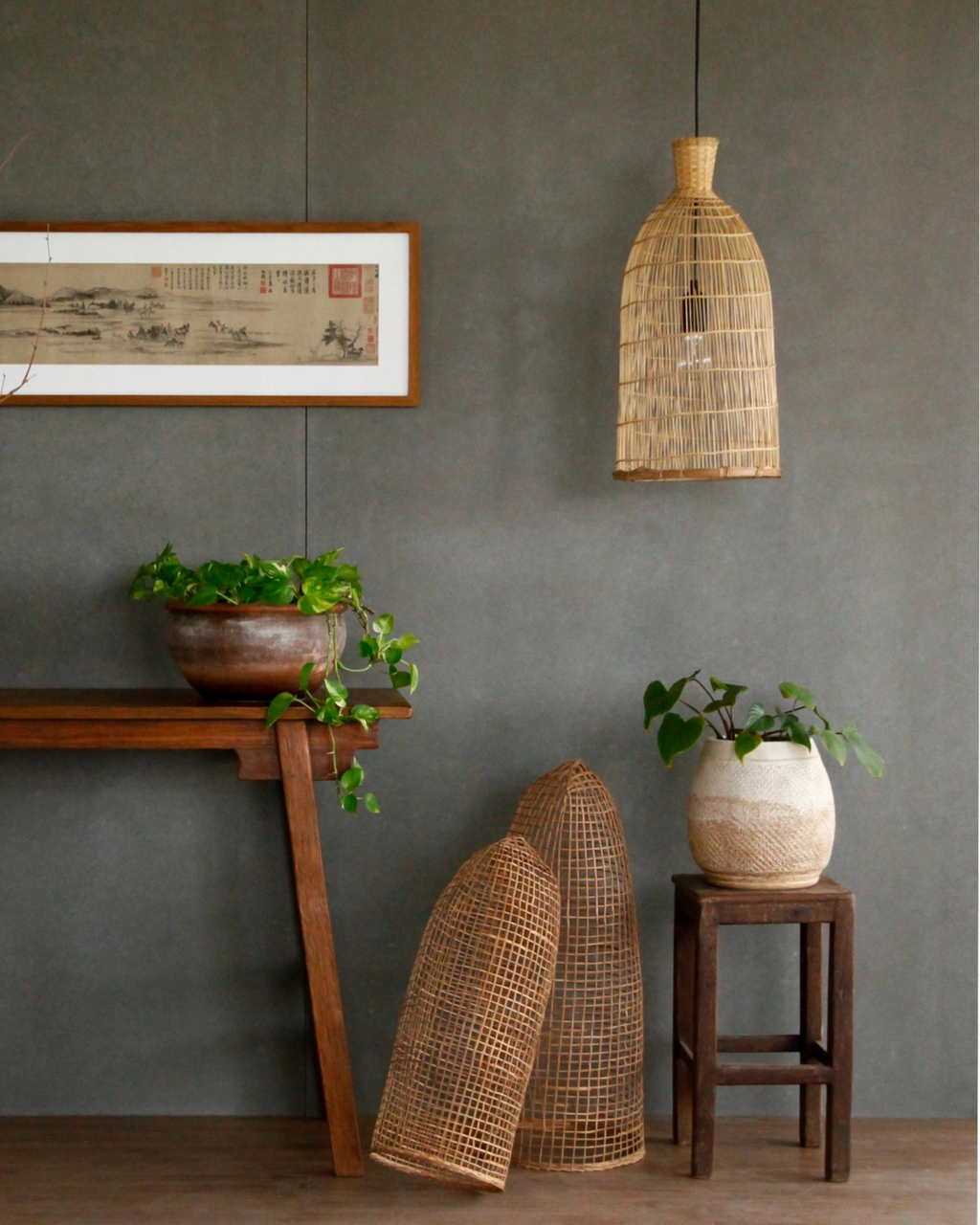 Decorative setting with a framed artwork, wooden table, plants, and hanging bamboo lamp against a gray wall.