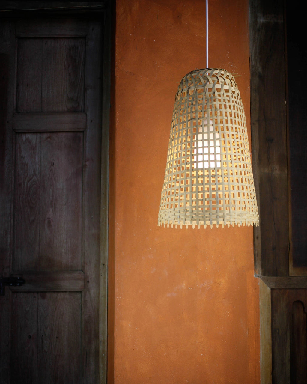 Woven bamboo pendant light hanging against a warm orange wall and a wooden door