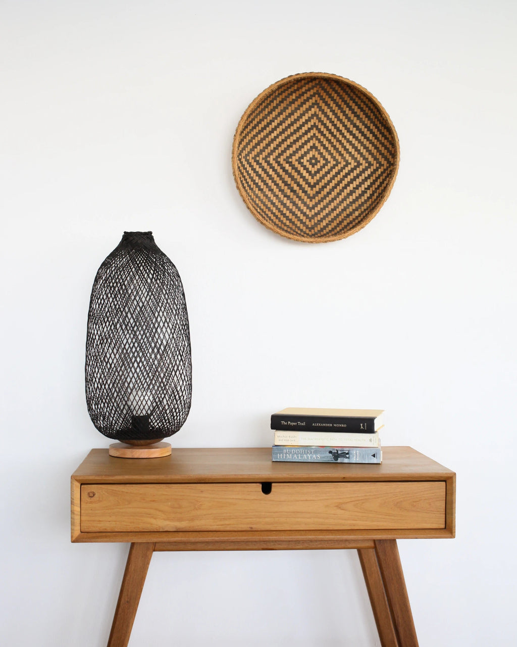 Wooden side table with a black woven bamboo lamp and books, featuring a woven basket on the wall above.