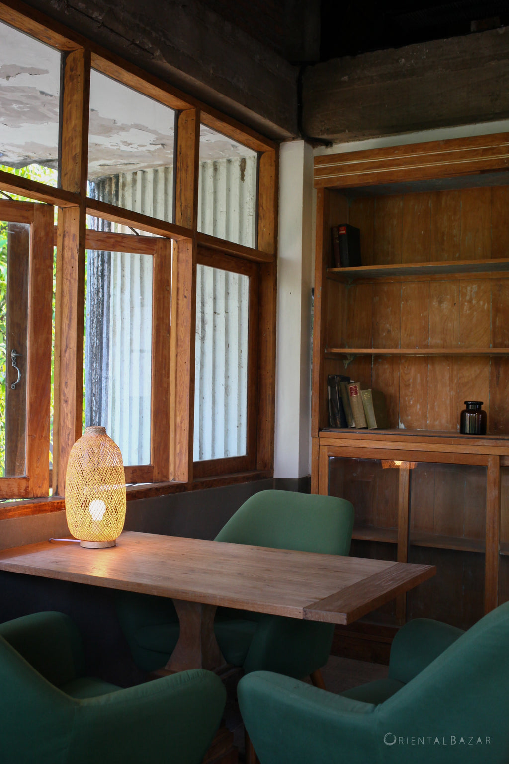 Organic Modern interior with a bamboo standing lamp on a wooden table next to a window, surrounded by green chairs, and a bookshelf.