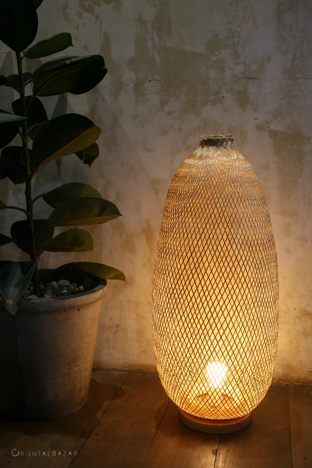 Decorative bamboo floor lamp with on a wooden floor next to a plant, against a textured wall.