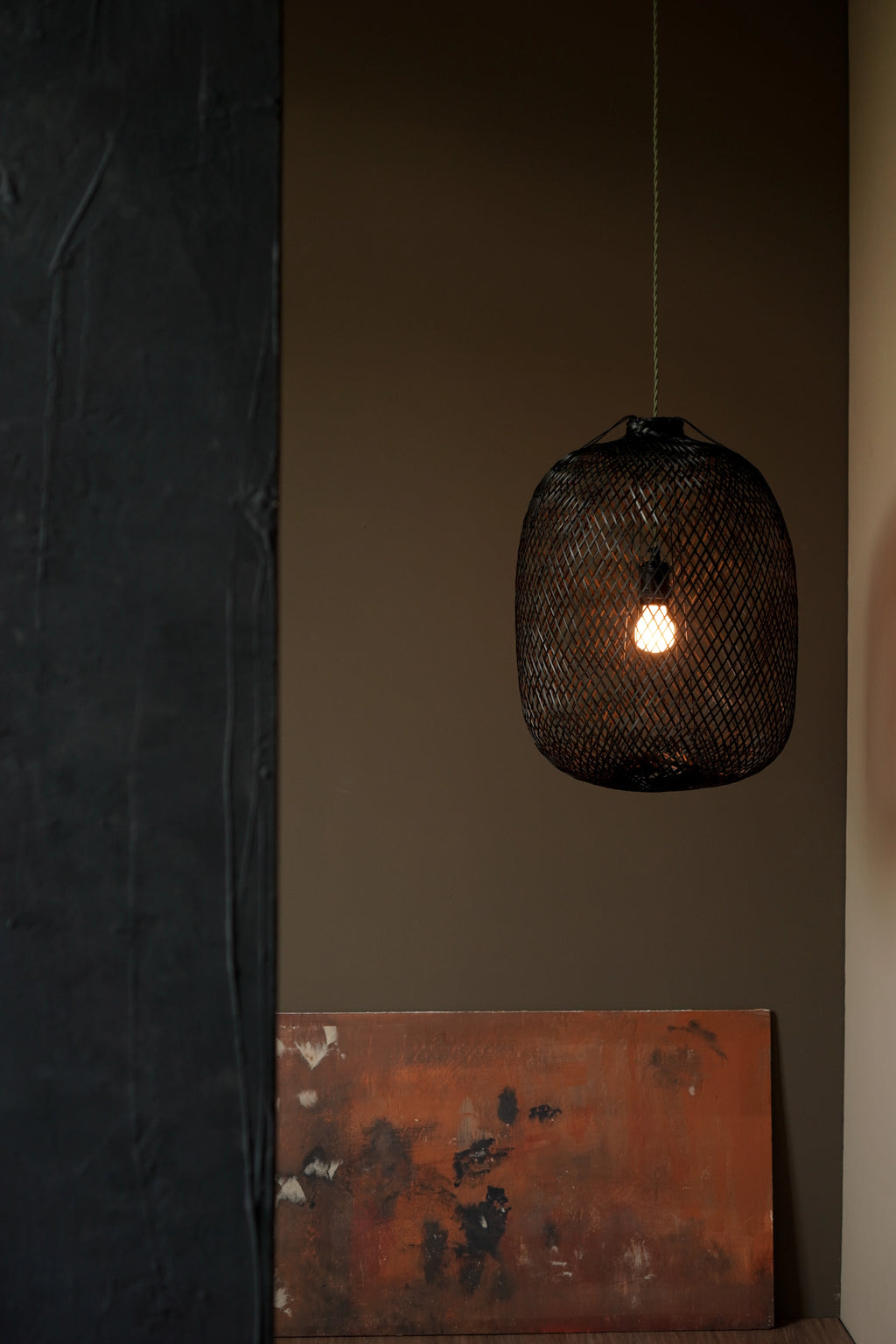 Black bamboo pendant light hanging against a dark wall with a textured surface and a warm colored art piece