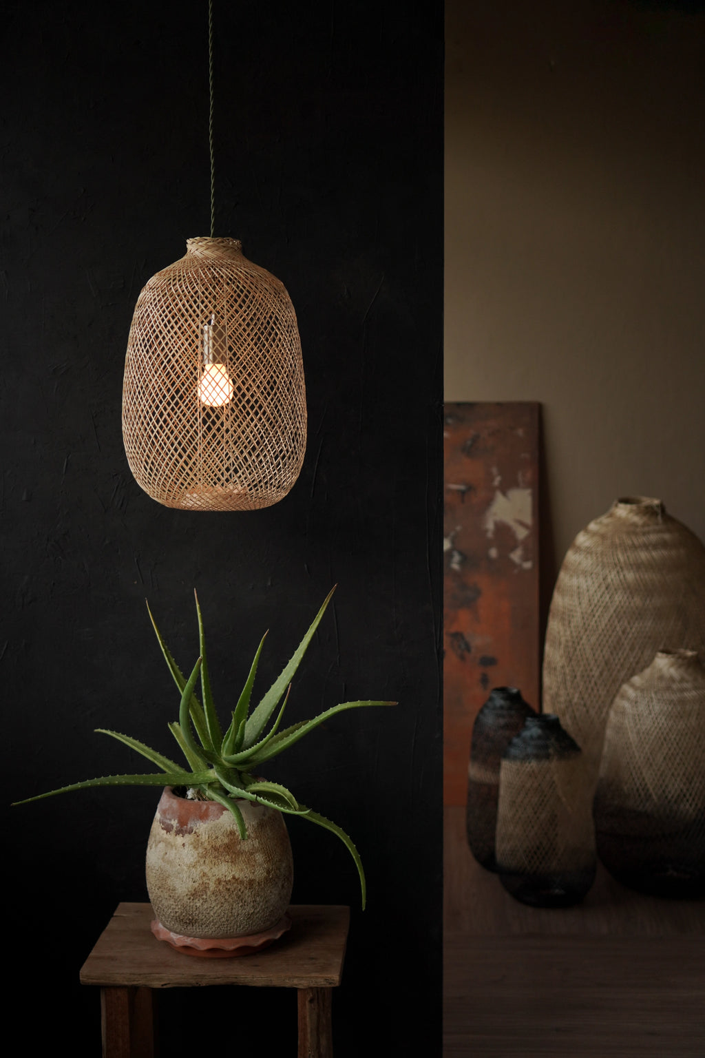 Woven bamboo pendant light fixture on a dark wall with a potted plant and additional woven bamboo lamps in the background.