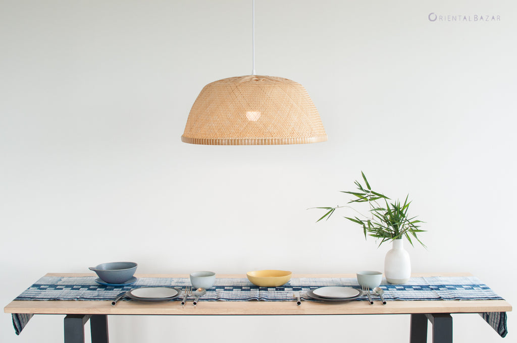 Dining table with a woven bamboo pendant light, plates, and a vase on a white background