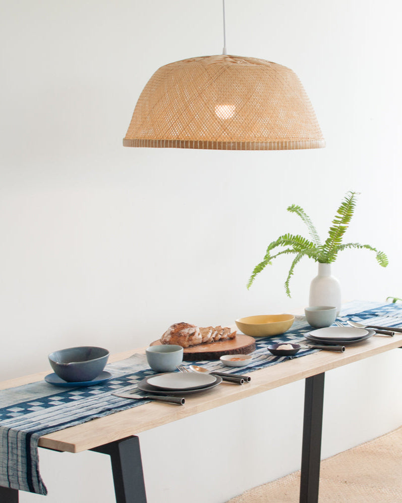 Dining table with a woven bamboo pendant light, plants, and a blue runner.