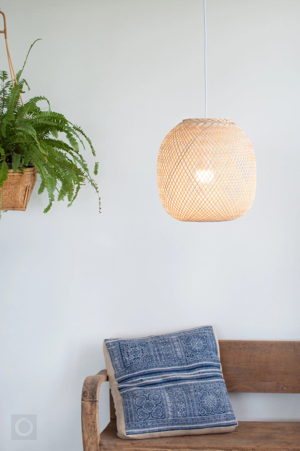 Woven bamboo pendant light hanging above a wooden bench with a blue cushion against a white wall next to a fern plant