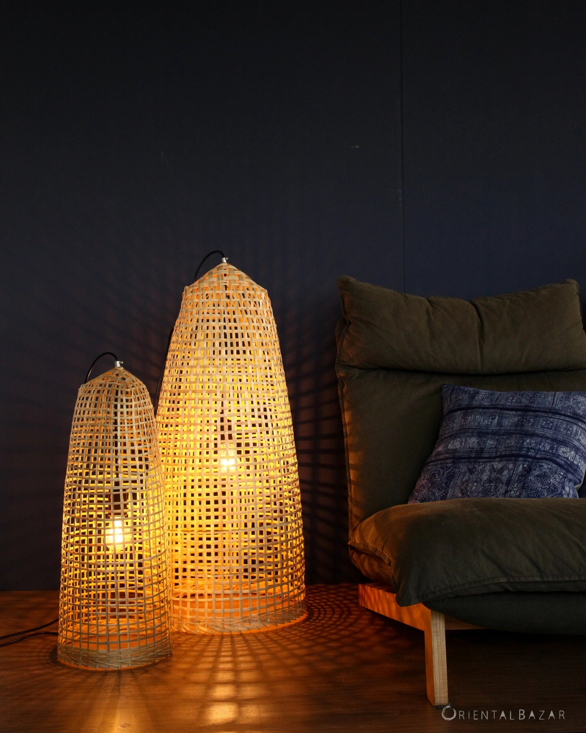 Woven bamboo standing lampshades on a wooden floor next to a sofa with a dark wall in the background