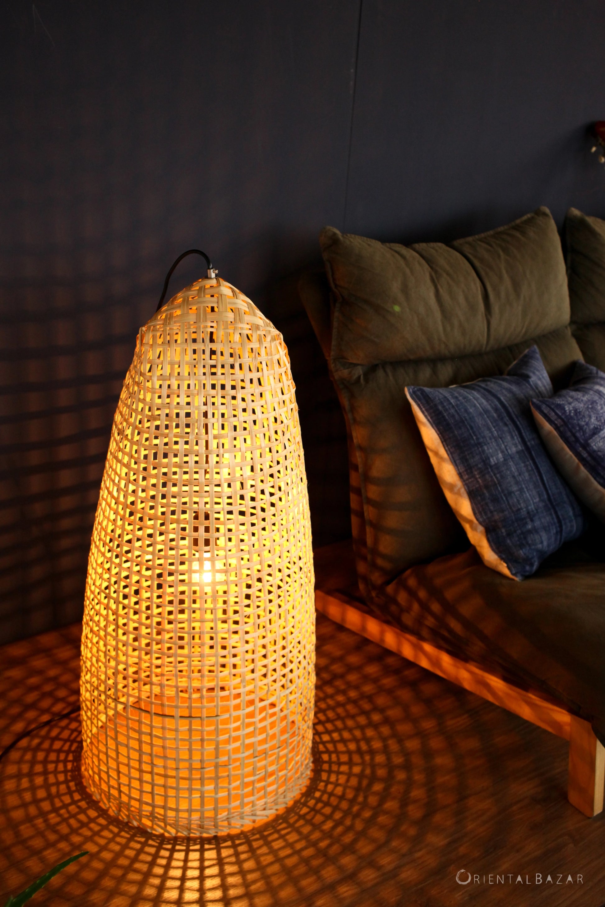 Woven bamboo standing lampshade casting a warm glow on a wooden floor with a dark wall in the background, next to a sofa